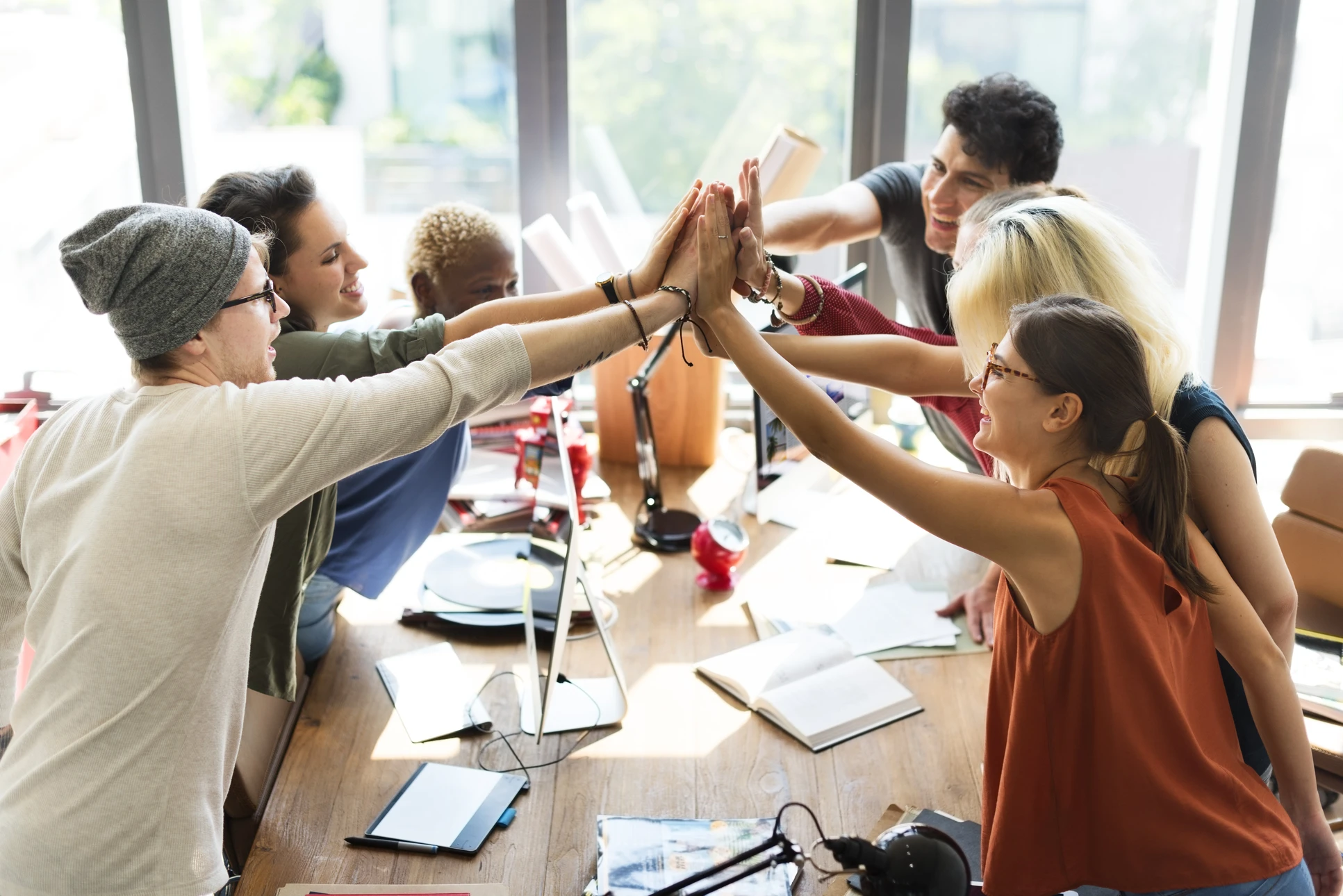 Groupe de six jeunes professionnels effectuant un high-five collectif autour d'une table de travail dans un bureau moderne