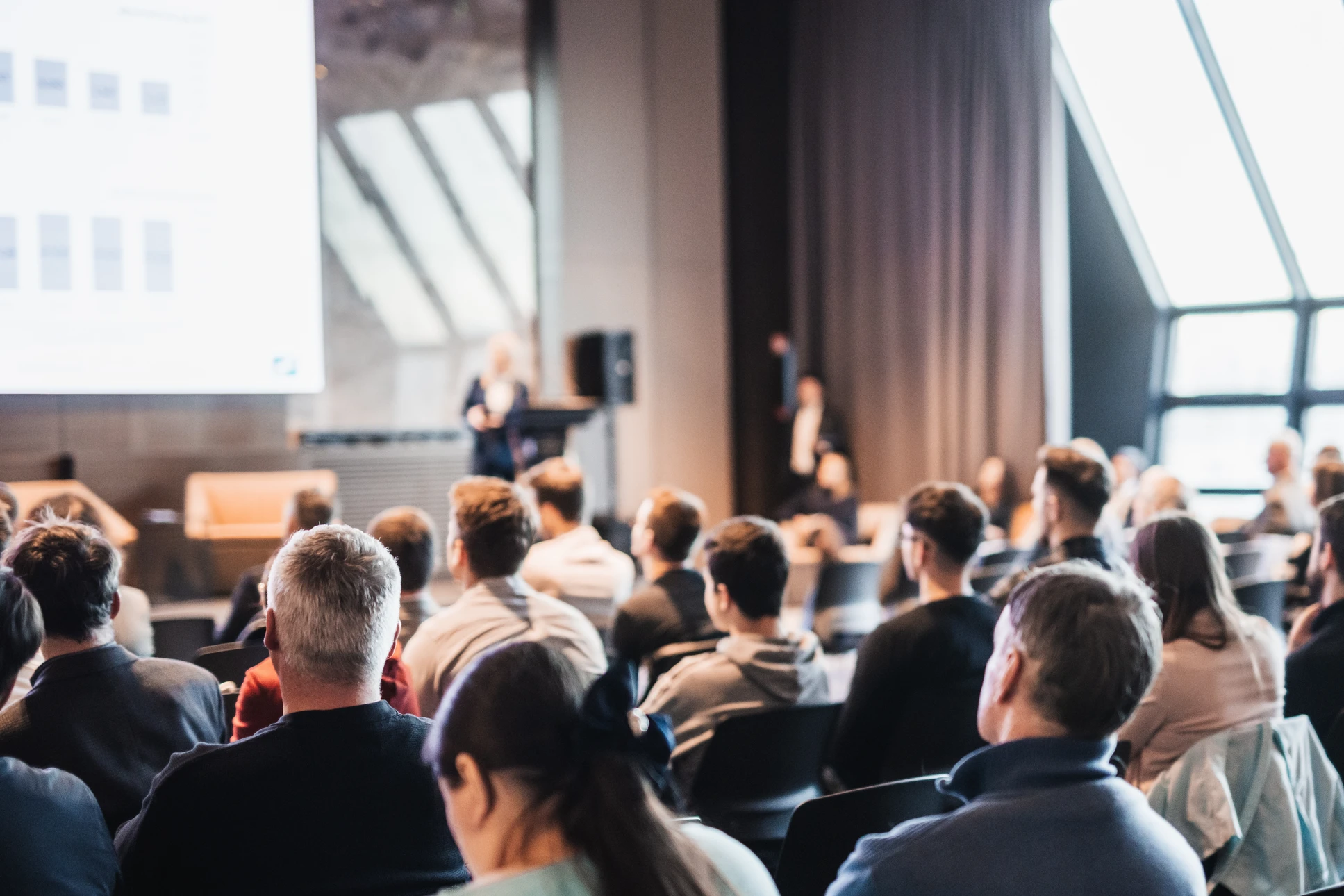 Salle de séminaire professionnelle spacieuse et élégante, avec public assis confortablement face à une scène bien éclairée. Un présentateur engageant anime la session devant l'audience attentive. La décoration est raffinée et professionnelle, avec un bon système de sonorisation et d'éclairage visible. L'atmosphère générale transmet professionnalisme, confiance et dynamique événementielle de qualité.